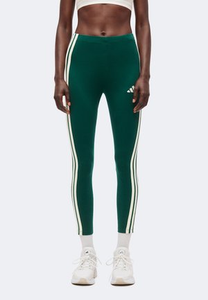 Atletisk person iført mørkegrønne leggings med hvide striber ned langs siderne, hvide sokker og hvide atletik sko.