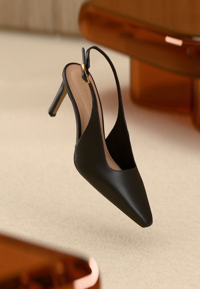 Mustat kärjellään terävät slingback-korkokengät ohuella remmillä ja kultaisella soljella beigen maton päällä ruskean kalusteen vieressä.