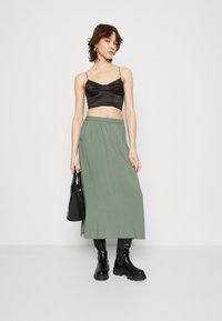 Top court en satin noir avec des fines bretelles, associé à une longue jupe verte fluide. Le modèle tient un sac noir et porte des bottes noires chunky.