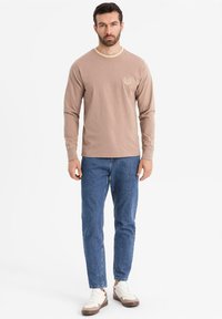 Beige Langarm-T-Shirt aus Baumwolle mit rundem Ausschnitt und dezentem Logo. Kombiniert mit blauen Denim-Jeans und braunen/weißen Sportschuhen.