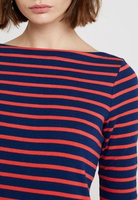 Tummansininen pitkähihaa paita, jossa on vaakasuoria punaisia raitoja. Ominaisuuksiin kuuluu leveä kaula-aukko ja pehmeä, joustava kangasosuus.