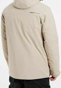 Veste beige isolante avec capuche, présentant une texture lisse, un design matelassé et un logo sur le haut du dos. Manches avec poignets ajustables.