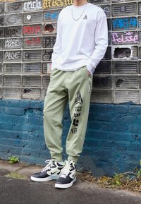 Weißes Langarmhemd mit Logo, grüne Sweatpants mit Druckdesigns und schwarze/gummierte High-Top-Sneaker mit bunten Akzenten.