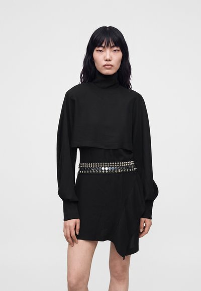 Femme aux longs cheveux noirs portant un haut noir à col montant et une jupe courte avec une ceinture cloutée, sur un fond gris uni.