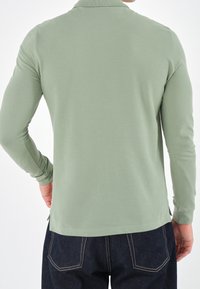 Langsleeve polo shirt, lichtgroene stof, zachte textuur, zijsplitten aan de onderrand, gecombineerd met donkere denim broek, gezien van achteren.