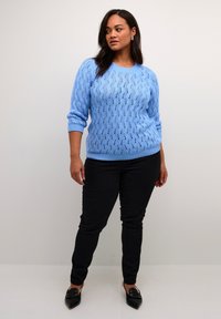Hellblaues Strickpullover mit einem strukturierten, spitzenähnlichen Muster, Rundhalsausschnitt und langen Ärmeln, kombiniert mit schwarzen Slim-Fit-Hosen und schwarzen Schuhen.