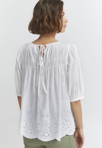 Blouse en coton blanc avec des manches bouffantes, un dos smocké et une fermeture à nouer. Présente de la broderie ajourée et des détails en dentelle au niveau de l'ourlet.