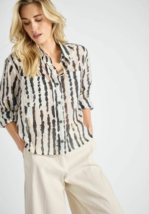 Blonde vrouw met los golvend haar, gekleed in een semi-transparant, patroonrijk overhemd met opgerolde mouwen en lichtgekleurde broek, handen in de zakken.