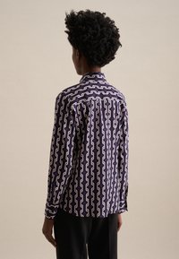 Camicia a maniche lunghe di colore viola con un motivo geometrico in crema. Presenta un colletto a punta e uno spacco sul retro con bottoni.
