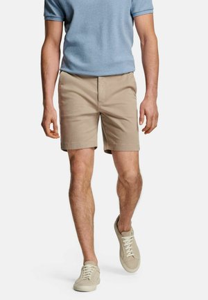 Hellbraune Shorts mit flachem Frontdesign, aus Baumwollmaterial, kombiniert mit einem blauen Kurzarmhemd und beigen Sneakern.