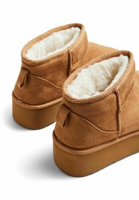 Botas de tobillo de ante marrón con forro de lana blanca y suela de goma plana. Presentan detalles de costura y una lengüeta trasera para facilitar su puesta.