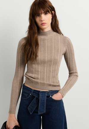 Donna con lunghi capelli castani che indossa un top beige a coste a maniche lunghe e jeans blu scuro a vita alta con cintura in denim annodata.