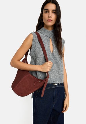 Kobieta ubrana w szary sweter bez rękawów i ciemne dżinsy, trzymająca dużą brązową zamszową torbę na ramię z paskiem i klamrą.