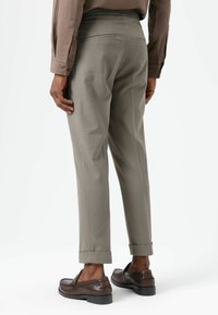 Lichtgrijze, op maat gemaakte broek met een taps toelopende pasvorm, voorzien van twee achterzakken en omgeslagen zomen, gecombineerd met donkerbruine instapschoenen.