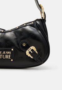 Czarna torebka z ekoskóry ze złotymi okuciami, ozdobiona ćwiekami, z odpinanym paskiem. Posiada logo na plakietce i charakterystyczne zapięcie.