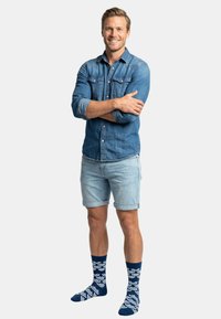 Camisa vaquera con las mangas remangadas, combinada con pantalones cortos azul claro. Calcetines azul marino con patrón de ondas blancas. Modelo de pie con los brazos cruzados, sonriendo.