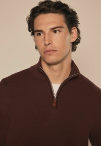Bordo zip-up pulover s rebrastom teksturom, sadrži srebrni zip i kožni naglasak na izrezu.