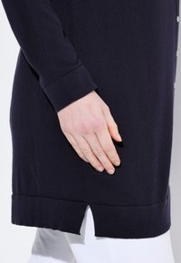 Mörkblå stickad tröja med ribbad struktur, långa ärmar och en sidoklyvning i fållen. Knappar synliga längs fram.