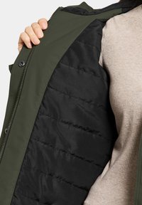 Groen buitenmateriaal met een zwarte gewatteerde voering; voorzien van een rits- en knoopsluiting. Het interieur heeft een soepele textuur.