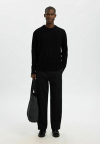 Černý pletený svetr s kulatým výstřihem, doplněný volnými černými kalhotami. Modelka drží tmavě šedou kabelku a má na sobě černé boty.