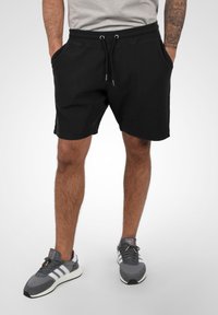 Shorts noirs en tissu texturé, avec taille élastique et cordon de serrage. Le modèle les associe à des baskets grises avec des rayures blanches et un logo.