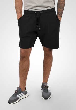 Shorts noirs en tissu texturé, avec taille élastique et cordon de serrage. Le modèle les associe à des baskets grises avec des rayures blanches et un logo.