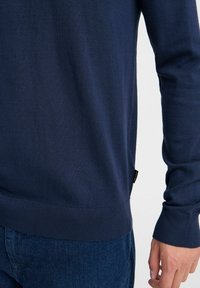 Marineblå, langermet strikket genser med ribbestrikket kant og mansjetter, med en liten logo-merkelapp på siden. Myk tekstur, figursydd design.