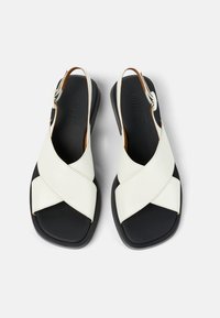 Valkoiset nahka-slingback-sandaalit, joissa on leveä ristikkäinen nauhamalli, musta kumipohja ja ruskeat yksityiskohdat sisäpuolella.