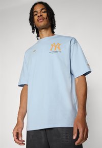 T-shirt en coton bleu clair avec un logo NY orange et le texte "ÉTABLI EN 1903, LE BRONX". Manches courtes, coupe décontractée, détails minimaux.