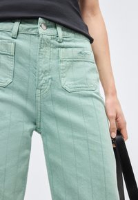 Κοντινό πλάνο ενός ατόμου που φοράει ανοιχτοπράσινο παντελόνι με κάθετες ραφές και τσέπες τύπου patch, κρατώντας ένα μαύρο δερμάτινο λουράκι στο δεξί του χέρι.