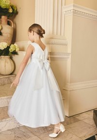 Junges Mädchen in weißem Kleid mit großer Schleife hinten, die nach unten zeigt, steht auf Marmortreppen neben großen Vasen mit grünen Blumen.