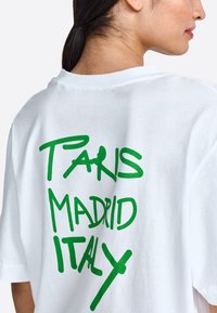 Balta medvilninė marškinė su pertekliniu kirpimu, ant nugaros ryškus žalias tekstas "PARIS MADRID ITALY". Paprastas dizainas ir sklandi tekstūra.