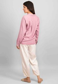 Chemise rose à manches longues avec une coupe décontractée, détail de dos patron, associée à un pantalon blanc ample et des sandales plates. Texture douce.