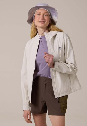 Jeune femme portant une veste beige clair, une chemise lavande, un short marron et un chapeau seau lavande assorti, souriante et tenant le cordon de sa veste.