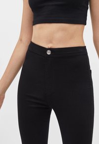 Černé úzké džíny z hladké elastické látky. Mají zapínání na knoflíky vpředu a vysoký pas. Žádné viditelné kapsy ani vzory.