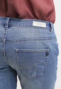 Jeans de mezclilla azul que presentan un bolsillo trasero visible con detalles de costura y una etiqueta de s.Oliver arriba. La tela muestra un ligero desgaste.