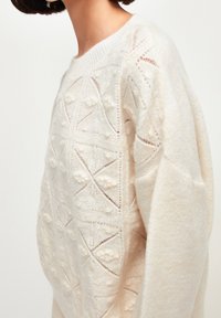 Maglione di colore crema con texture, caratterizzato da motivi geometrici, dettagli a maglia e una vestibilità morbida con scollatura rotonda.