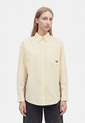 Un mannequin porte une chemise boutonnée jaune pâle avec une poche poitrine et des bandes bordeaux sur les côtés, associée à un jean foncé, sur un fond uni.