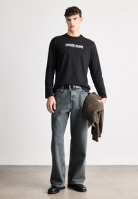 Camisola preta de manga longa com o logótipo branco "CALVIN KLEIN", combinada com jeans de pernas largas azul claro e uma jaqueta xadrez castanha. Sapatos pretos.