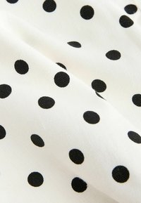 Tissu blanc avec une surface légèrement texturée présentant un motif de pois noirs répartis de manière régulière et de plis doux.