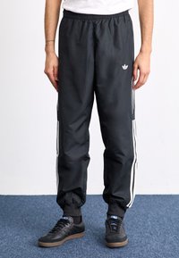 Μαύρες φόρμες με ελαστική μέση, με λευκές πλάγιες ρίγες. Το υλικό φαίνεται λείο με ελαφριά γυαλάδα; οι μανσέτες είναι μαζεμένες στους αστραγάλους.