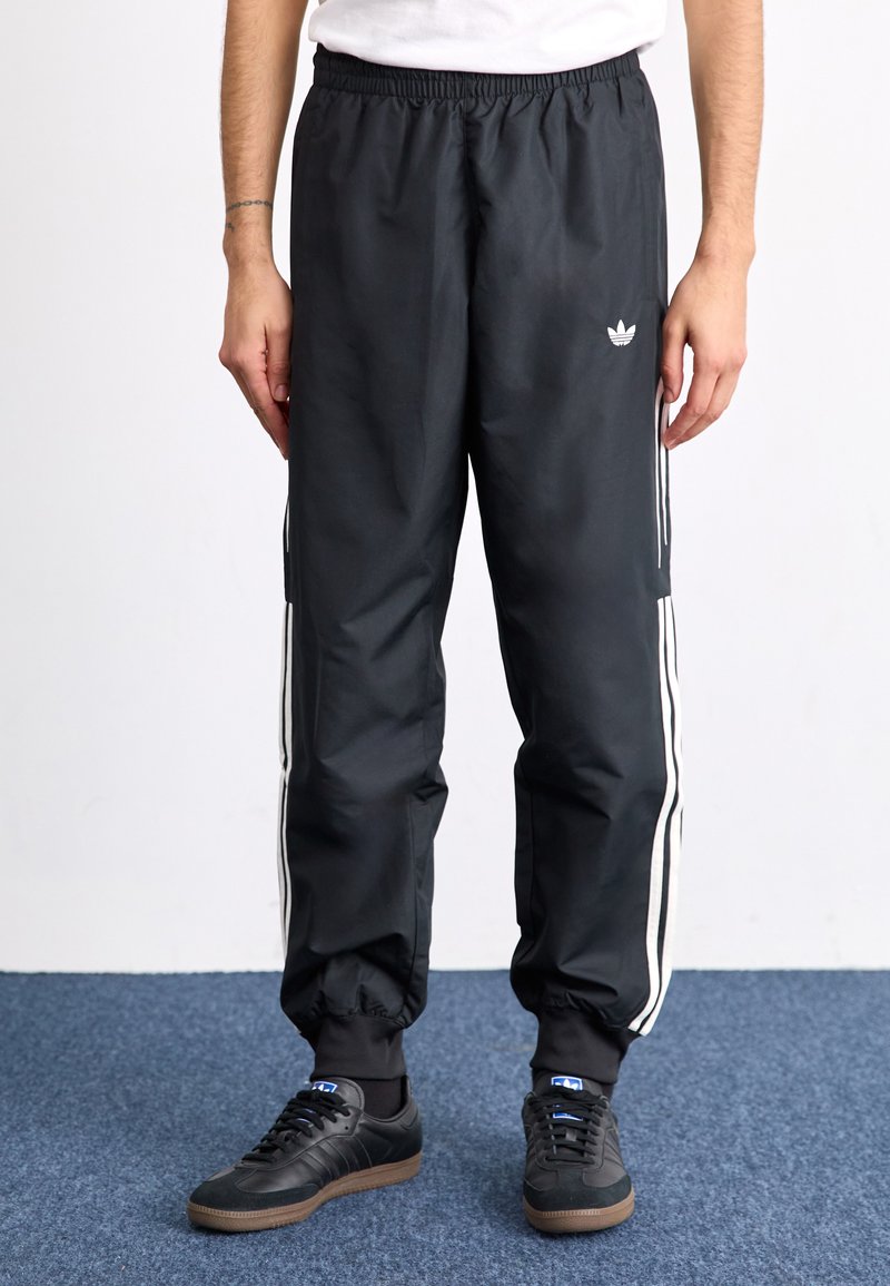 Μαύρες φόρμες με ελαστική μέση, με λευκές πλάγιες ρίγες. Το υλικό φαίνεται λείο με ελαφριά γυαλάδα; οι μανσέτες είναι μαζεμένες στους αστραγάλους.