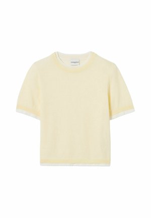 Jeune femme debout portant un haut jaune clair à manches courtes, un pantalon large bleu à motifs, des chaussures marron et portant un sac blanc à l'épaule.