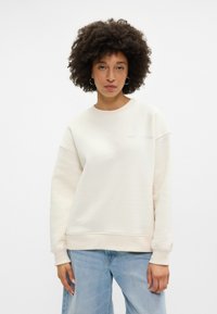 Krémová mikina vyrobená z měkkého materiálu, s uvolněným střihem, klasickým výstřihem a jemnou výšivkou na hrudi.