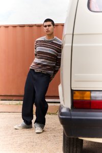 Pruhovaný pletený svetr v hnědých a modrých tónech, tmavé džíny a šedé boty. Model se opírá o bílý dodávkový vůz s výraznými zadními světly.