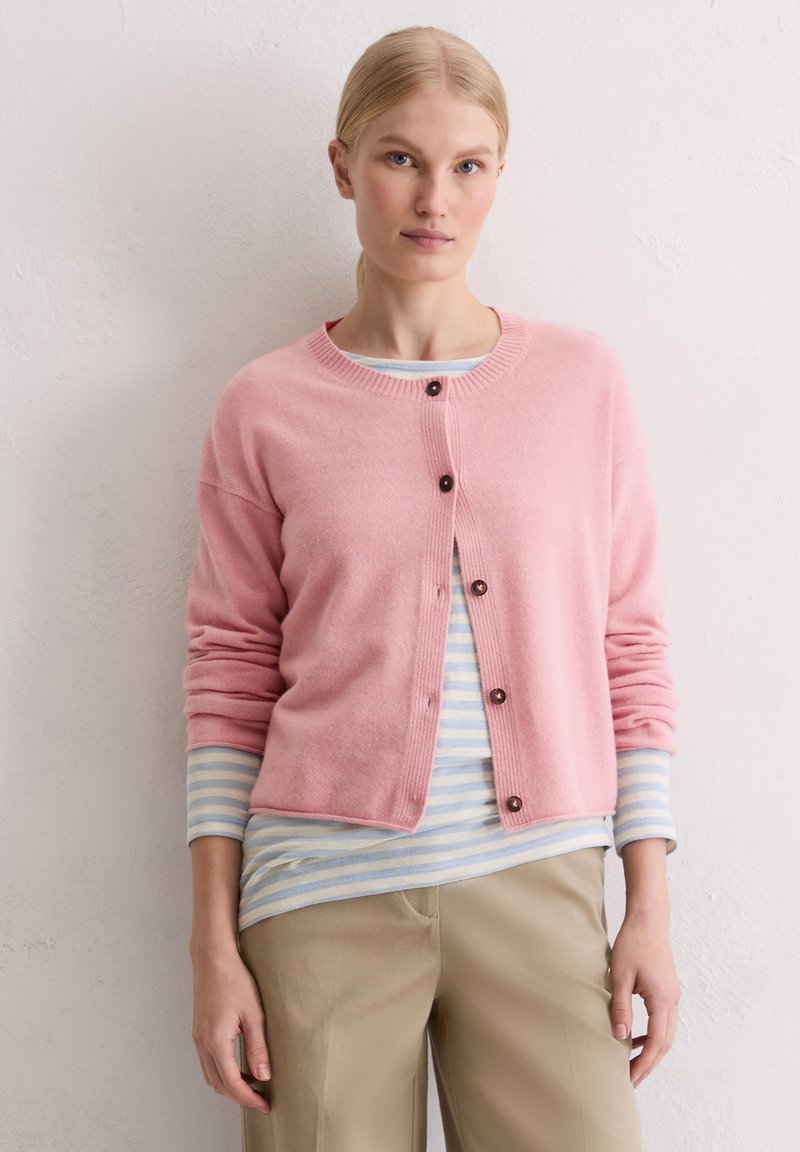 Gilet en cachemire rose avec un col rond et des boutons, associé à une chemise à manches longues rayée bleue claire et blanche et un pantalon beige.