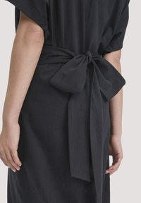 Frau in einem schwarzen Kleid mit kurzen Ärmeln, von hinten gezeigt, mit einer großen gebundenen Schleife um die Taille.