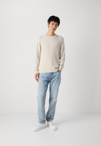 Beżowy sweter z plecionki, z prążkowanymi mankietami, w połączeniu z jasnoniebieskimi dżinsami i białymi sneakersami. Model stoi na jednolitym tle.