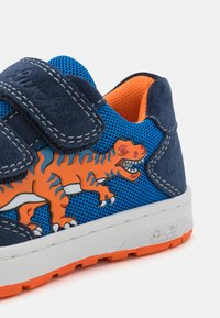 Μπλε δίκτυο αθλητικού παπουτσιού με ναυτικό σουέτ λεπτομέρειες, διαθέτει γραφικό με πορτοκαλί δεινόσαυρο, ρυθμιζόμενο ιμάντα Velcro και λευκή καουτσούκ σόλα με πορτοκαλί εξωτερική σόλα.