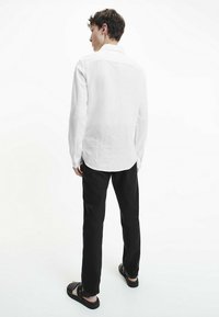Camicia bianca in lino a maniche lunghe con colletto a punta, orlo curvo, polsini con bottoni, indossata con pantaloni neri e sandali. Tessuto morbido e traspirante.